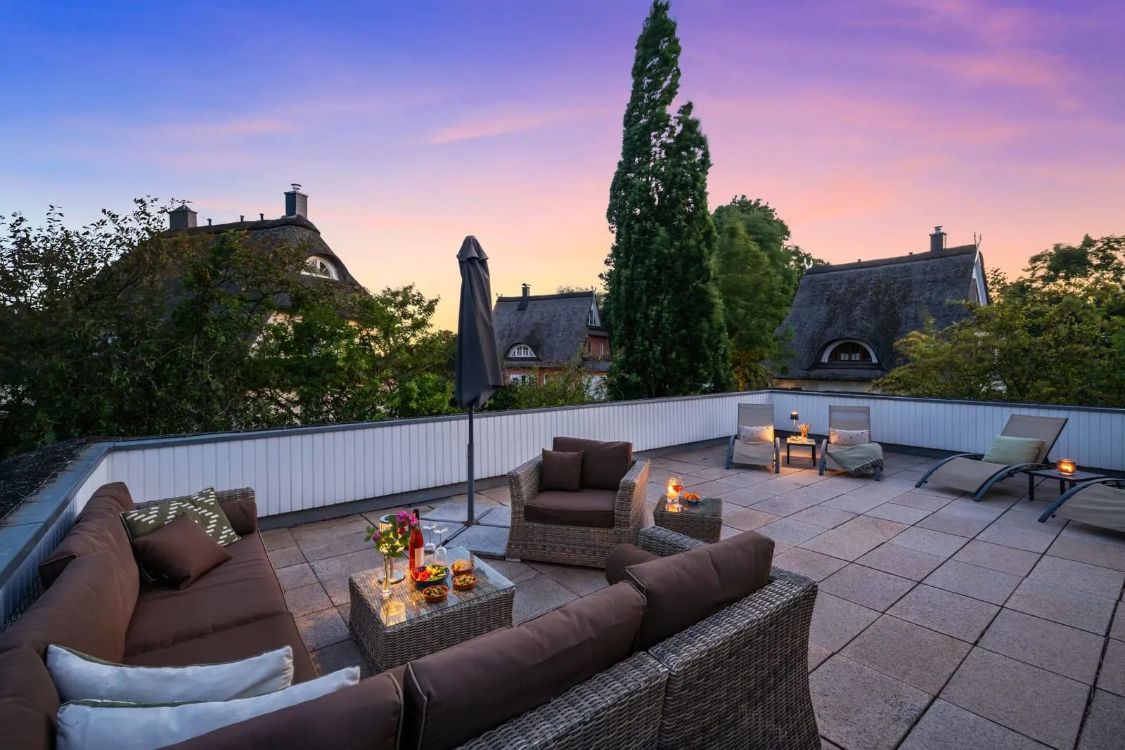 Dieselbe Dachterrasse nach dem Styling – gepolsterte Lounge-Möbel, gedeckter Tisch mit Blumen und Kerzen in warmer Abendstimmung