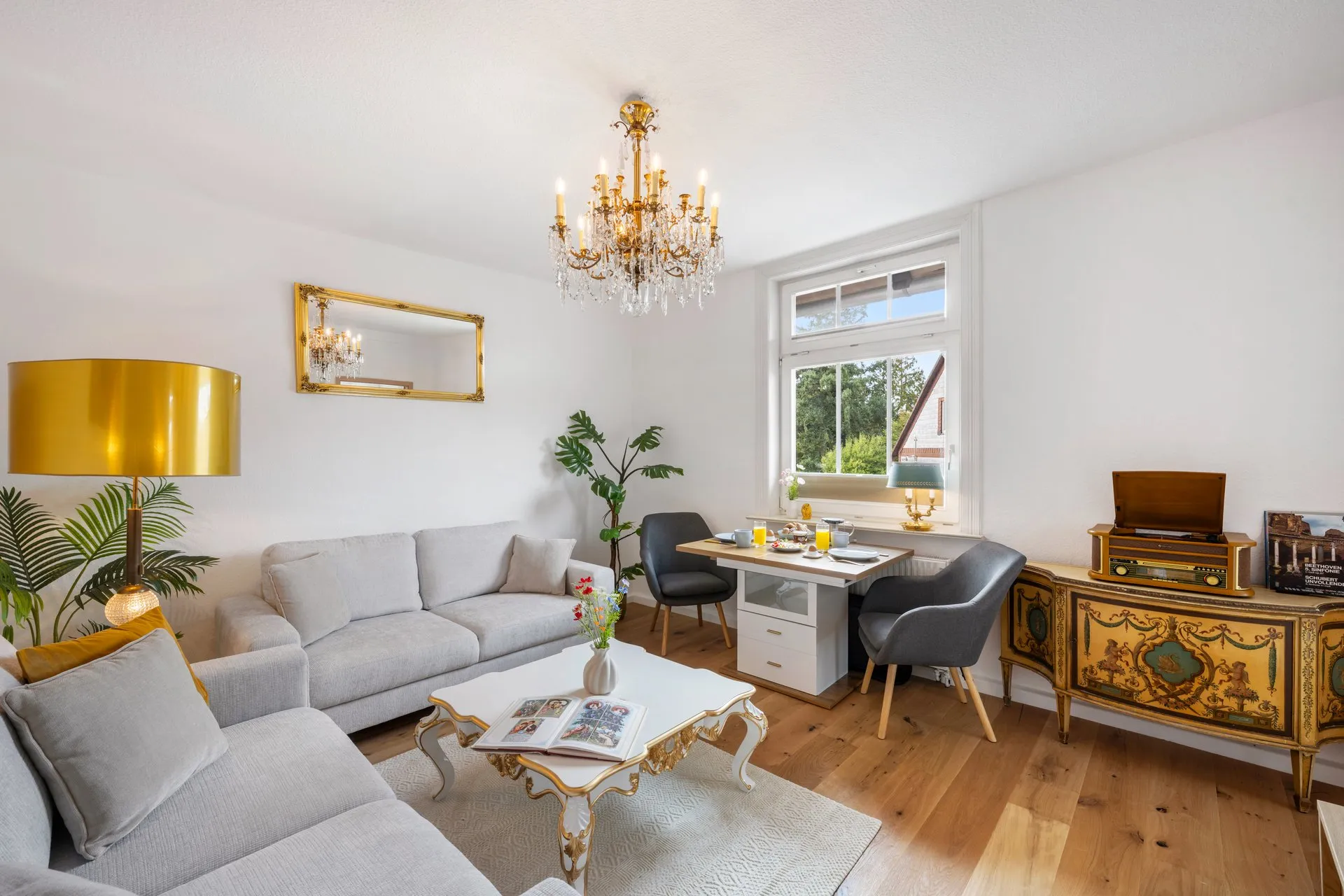Wohnraum mit Kristallleuchter, grauen Sofas und verziertem Sideboard, am Fenster gedeckter Tisch