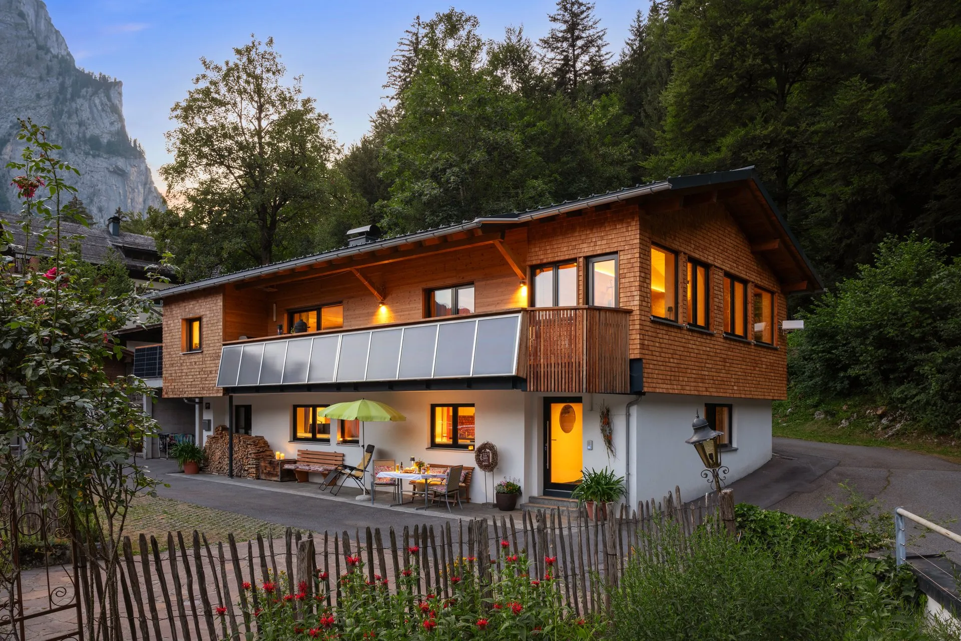 Holzchalet in der Abenddämmerung, warm erleuchtete Fenster, davor eine Terrasse mit Sonnenschirm