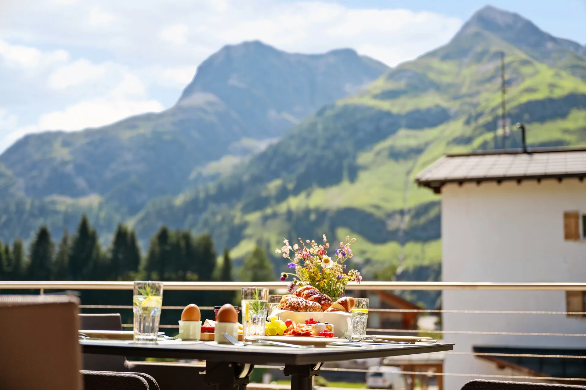 Gedeckter Frühstückstisch auf dem Balkon mit Hefezopf und Wildblumen, dahinter grüne Berggipfel
